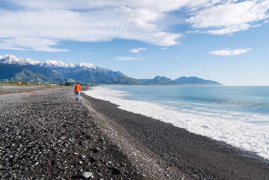 A man walks along a rocky beach with a stunning view of snow-capped mountains and the ocean.