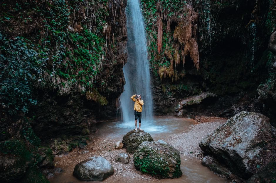 A lone adventurer stands on rocks near a waterfall in Seydikemer, Turkey, surrounded by lush greenery.