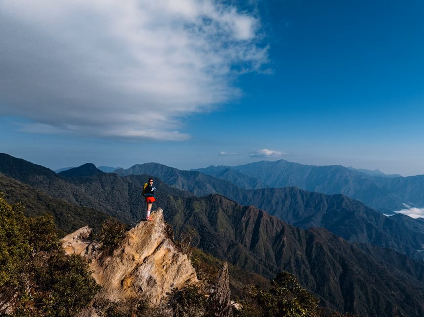 Hiker stands atop a rocky cliff overlooking expansive mountain range in Vietnam.