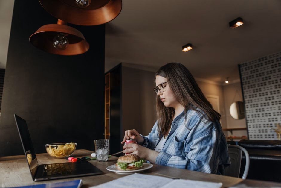 Woman eating lunch at home while working on laptop, featuring chips and a burger on a wooden table.