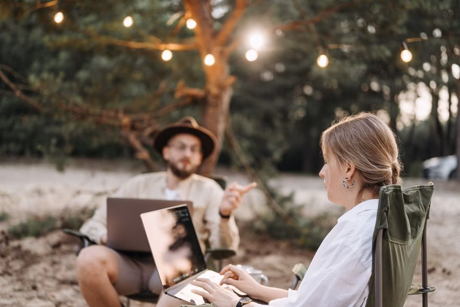 Two adults working on laptops in a rustic outdoor setting with ambient lighting.