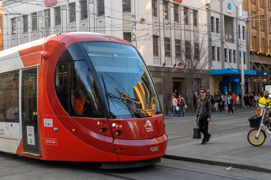 A vibrant red tram on a busy street in Loftus, NSW, Australia, with pedestrians and cyclists.