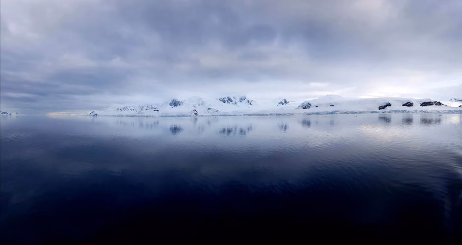Calm waters reflecting Antarctic mountain range beneath a cloudy sky.