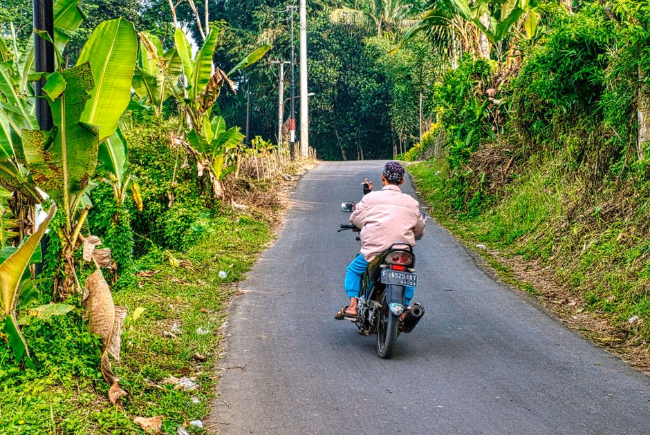 A motorcyclist navigates a rural road in West Java, surrounded by green vegetation.
