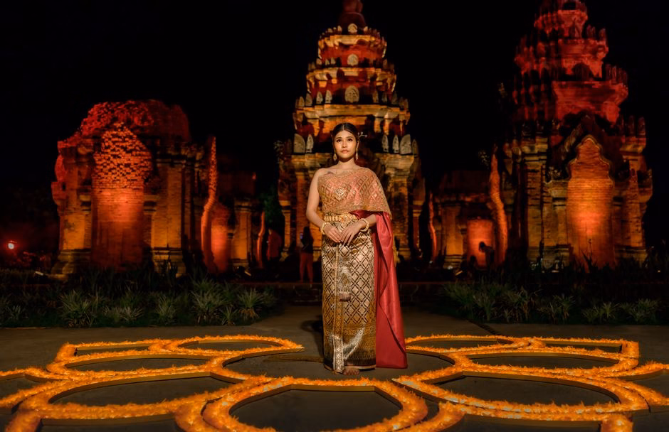 A woman in traditional attire stands elegantly at an illuminated ancient temple adorned with candles at night.