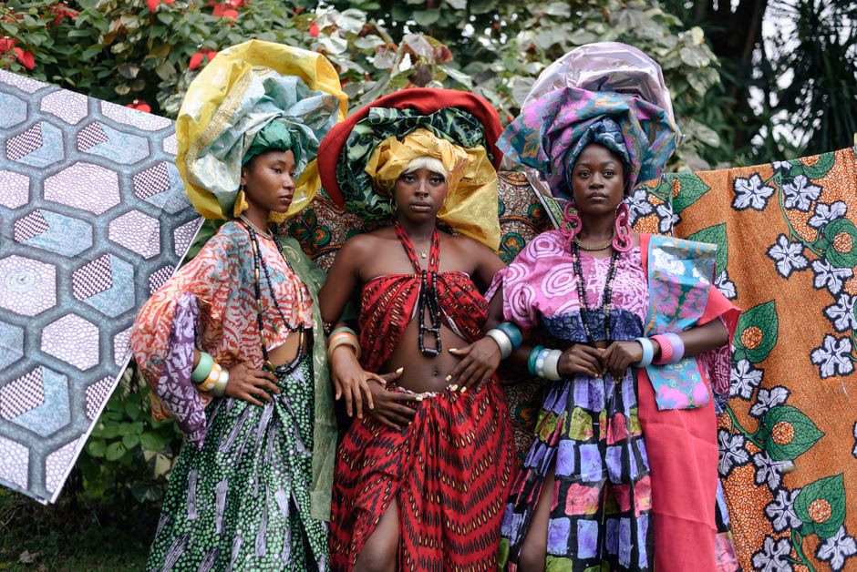 Three women in colorful Nigerian attire with headwraps, posing confidently outdoors.